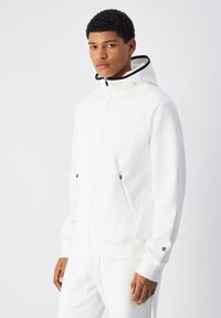 Felpa bianca con zip e cappuccio foderato, dettagli neri, due tasche laterali e maniche con polsini, con dettaglio del logo sul polso.