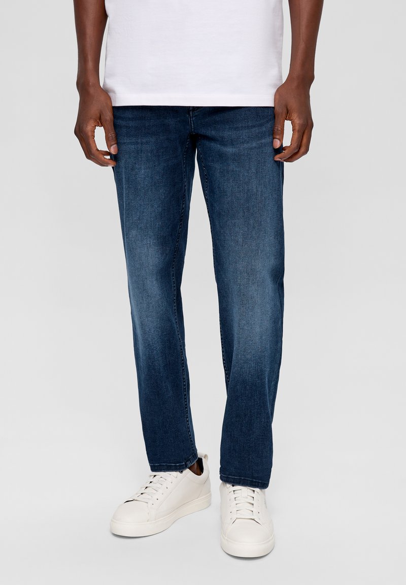 Dunkelblaue Jeans im geraden Schnitt mit glatter Textur, getragen mit weißen flachen Sneakers. Ein schlichtes weißes Hemd ist am Saum sichtbar.