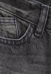 Zbliżenie na kieszeń czarnych dżinsów z widocznym niebieskim przeszyciem i detalem pętli na pasek.