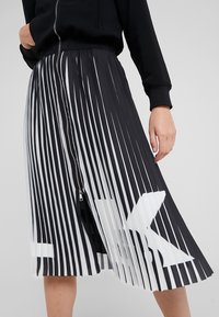 Schwarz-weißer plissierter Rock mit vertikalen Streifen, ausgestattet mit einem Reißverschluss und einem großen weißen grafischen Detail am Saum.