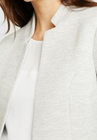 Veste gris clair avec un col structuré, une texture en chevrons et une silhouette ajustée, associée à un haut blanc en dessous.