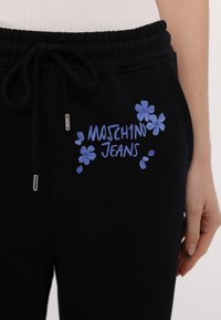 Mustat verkkarit, joissa on naru vyötäröllä, sinisillä kukkakuvioilla ja "MOSCHINO JEANS" -teksti vasemmassa lahkeessa.
