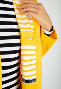Veste jaune avec des rayures beiges et jaunes contrastantes à l'intérieur. Présente une poche zippée et un haut à rayures noir et blanc en dessous.