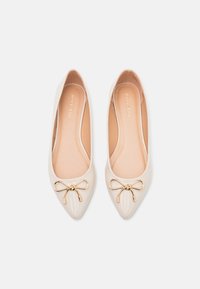 Paire de ballerines beige à bout pointu avec des accents de nœud doré sur le bout, présentées côte à côte sur un fond blanc.