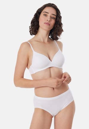 Kvinde med krøllet mørkt hår iført en hvid bh og hvide højtaljede trusser, stående mod en ensfarvet lys baggrund.