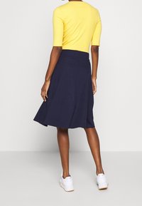 Keltainen ribatty toppi yhdistettynä tummansiniseen levenevään hameeseen. Hameella on sileä pinta ja se ulottuu polven alapuolelle, ja sen kanssa on valkoiset lenkkarit.