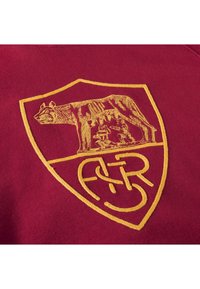 Bordeaux textiel met een geborduurd gouden logo. Het logo bevat een schildvorm met een beer en drie figuren aan de basis, met ingewikkelde details.