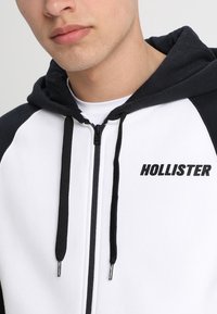 Hooded zip-up sweatshirt med en färgblockdesign i vitt och mörkblått, med svarta dragsnören och "HOLLISTER"-logotyp i fet text.