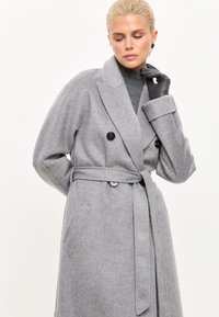Mantou de lână gri cu un design cu dublă închidere, nasturi mari negri, o curea asortată și buzunare laterale. Textură moale și croială lejeră.