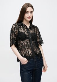 Sort blondebluse med knaplukning foran, korte ærmer og krave, med blomsterdesign og transparent struktur. Kombineret med jeans.