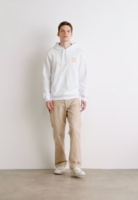 Felpa bianca con logo "HUGO" arancione, abbinata a pantaloni beige e sneaker bianche. Il modello è in piedi contro uno sfondo chiaro.