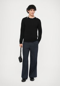 Zwarte gebreide trui met een ronde hals, gecombineerd met marineblauwe broek met streepjes en wijde pijpen. Houdt een kleine zwarte tas vast, staande op een effen achtergrond.