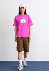 En model går fremad iført en lys pink t-shirt med grafik, brune shorts og en hvid kasket, imod en ensfarvet hvid baggrund.