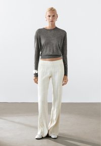 Grauer gestrickter Pullover mit einem cropped Design, kombiniert mit cremefarbenen weiten Hosen mit dezenten Streifen und weißen Slippers.