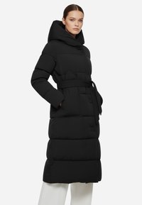 Lungo cappotto piumino nero con colletto alto, vita aderente e texture trapuntata. Presenta un ampio cappuccio e tasche laterali.