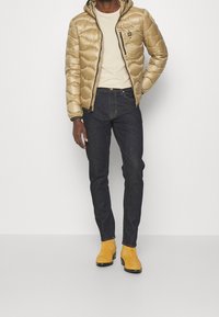 Giacca imbottita dorata con cappuccio, camicia crema, jeans scuri e stivaletti in camoscio giallo. Presenta dettagli con cerniera e tasche.