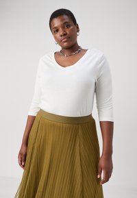 Top branco de gola em V e mangas longas, combinado com uma saia pregueada em verde-azeitona com um cós largo e detalhes de nervuras verticais. Colar de corrente em prata.