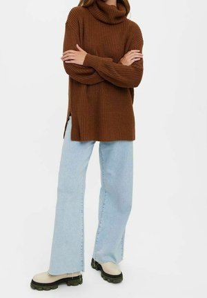 Femme portant un pull à col roulé marron surdimensionné, un jean large bleu clair et des bottes à semelle épaisse beige, les bras croisés.