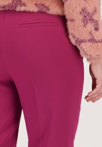 Pantalons taillés fuchsia avec une texture lisse, dotés d'une poche arrière et d'une coupe slim. Main posée sur la hanche.