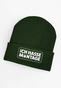 Dark green knit beanie with ribbed texture. Features a white patch with the text “¡CH HASSE MONTAGE!” and a cartoon character.
