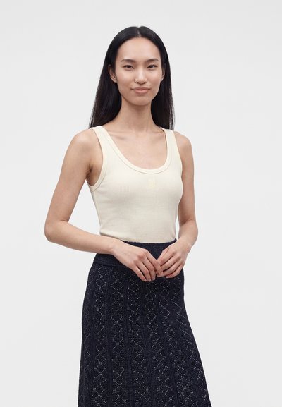 Femme aux longs cheveux lisses portant un débardeur sans manches crème côtelé et une jupe bleu foncé à motifs, debout devant un fond uni clair.