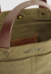 Detailní záběr na olivově zelenou texturovanou plátěnou tašku Levi's s hnědými koženými úchyty a viditelnou vnitřní etiketou Levi Strauss & Co.