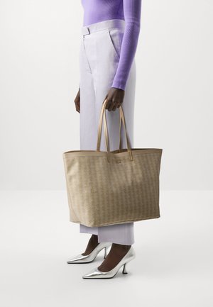 Sac fourre-tout beige avec un motif texturé. Il possède deux poignées robustes et une forme structurée. Porté avec un pantalon violet clair et des talons argentés.
