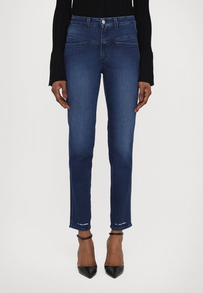 Mørkeblå denim jeans med slim fit, fem-lomme design og synlige sømme ved kanten. Bæres med sorte højhælede sko.