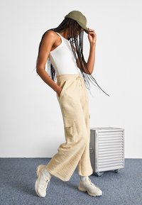 Beige teksturerte cargo-bukser med lommer, kombinert med en hvit tettsittende tank tops og en olivengrønn caps, fullført med lyse treningssko.