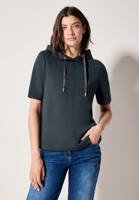 Sudadera corta de manga corta en verde oscuro con un bolsillo frontal, cordón alrededor del cuello y textura suave. Combinada con jeans de mezclilla azul.