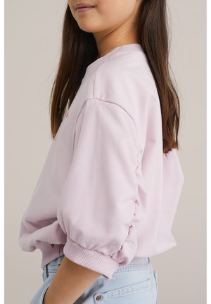 Jauna sieviete ar gariem tumšiem matiem, valkājot gaiši rozā džemperi ar pumpainām piedurknēm un gaiši zilas džinsa bikses, stāv sāniski pret neitrālu fonu.