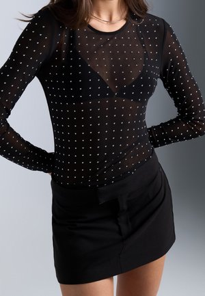 Femme portant un top noir transparent à manches longues avec de petits pois blancs sur un soutien-gorge noir, associé à une mini-jupe noire.