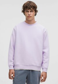 Sweatshirt lilas clair en tissu doux, avec un col rond et des poignets côtelés. Porté sur un t-shirt blanc à col rond.