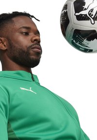 Groene sportjack met een hoge kraag en rits, met een wit Puma-logo. Een zwart-witte voetbalk rijdt erboven.