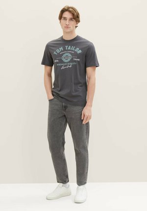 Graues Kurzarm-T-Shirt mit türkisfarbenem grafischen Text, kombiniert mit schwarzen Jeans und weißen Sneakers. Das Modell steht vor einem neutralen Hintergrund.