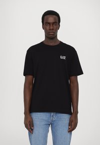 Svart bomull t-skjorte med korte ermer, rund hals og en liten hvit EA7-logo på venstre bryst. Båret med lyseblå jeans.