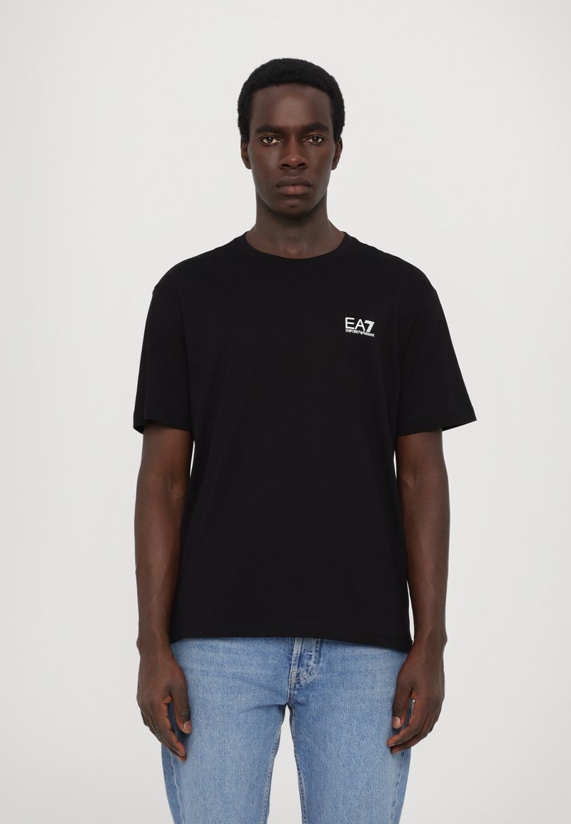 Svart bomull t-skjorte med korte ermer, rund hals og en liten hvit EA7-logo på venstre bryst. Båret med lyseblå jeans.