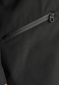 Jachetă neagră cu un buzunar orizontal cu fermoar și un mic fir de tragere, textură de țesătură netedă și detaliu ajustat la mansete.