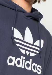 Sudadera con capucha navy con un gran logo blanco de Adidas que presenta tres hojas y tres franjas horizontales. Cuerda ajustable en la capucha.