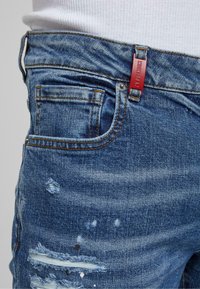Jeans de mezclilla azul claro con detalles desgastados, incluyendo parches deshilachados y manchas de pintura. Presenta una etiqueta de tela roja y bolsillos estándar.