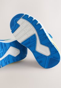 Semelles de deux baskets blanches avec des bandes de caoutchouc bleu montrant des motifs d'adhérence texturés sur un fond uni clair.
