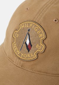 Brown cotton cap featuring an embroidered logo patch with "HILFIGER NEW YORK," a flag design in red, white, and blue, and textured fabric.