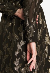 Robe noire avec broderie florale dorée, manches transparentes, détails de poignets à boutons, tissu texturé, vue en gros plan de la main et du vêtement.