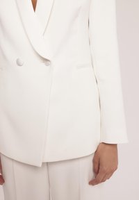 Biały żakiet dopasowany o podwójnym zapięciu, gładki materiał, zaokrąglone guziki oraz elegancka, strukturalna sylwetka.