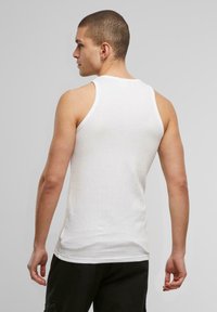 Weißes Baumwoll-Tanktop mit gerippter Textur, Rundhalsausschnitt und breiten Trägern, von hinten gezeigt. Schwarze Hose ist sichtbar.
