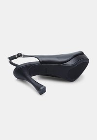 Crna kožna cipela na visoku petu sa otvorenom petom, glatke površine, zaobljenog vrha i teksturiranom potplatu. Sadrži podesivu traku i blok petu.