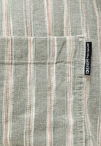 Κοντινή λήψη γκρι ριγέ υφάσματος με υφή ύφανσης και μια μαύρη ετικέτα "DENIM TOM TAILOR" ραμμένη στη δεξιά πλευρά.