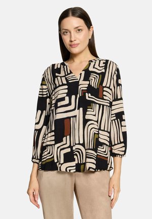 Kvinde iført en sort top med beige, brune og olivengrønne abstrakte geometriske mønstre og beige bukser, stående mod en ensfarvet baggrund.