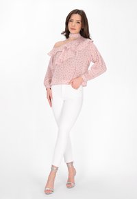 Rosa Bluse mit Pünktchenmuster und Rüschenverzierungen, Ein-Schulter-Design und langen Ärmeln, kombiniert mit weißen eng anliegenden Jeans und silbernen Riemchen-Heels.