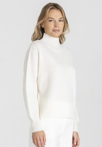 Biały dziergany sweter z golfem, z prążkowanymi mankietami i dołem. Miękka tekstura, luźny krój, lekko przylegający. Czysty, minimalistyczny wygląd.